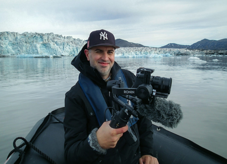mężczyzna w średnim wieku siedzący w motorówce typu rib, w dłoniach ma kamerę, do okoła jest woda a w tle znajdują się lodowce