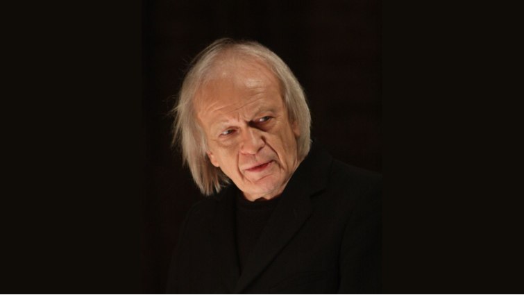 starszy mężczyzna, w dłuższych siwych włosach, ubrany w czarny garnitur