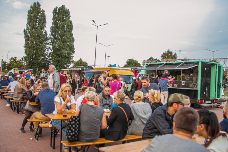 festiwal food trucków, ludzie siedzący przy stołach na placu, do okoła rozstawione są samochody z których serwowane są dania