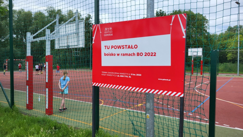 tablica informująca o tym że boisko powstało w ramach budżetu obywatelskiego 2022, w tle dzieci grające na boisku