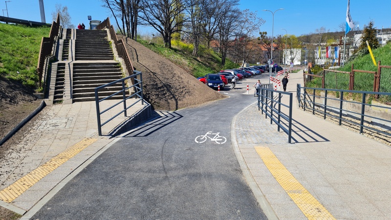 Na obrazie znajduje się droga rowerowa, wysokie schody z podjazdem na wózki, chodnik dla pieszych