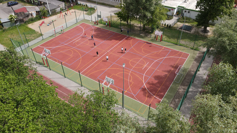 widok z lotu ptaka na nowe boisko z czerwoną nawierzchnią gumową na środku otoczoną zieloną gumową nawierzchnią, na boisku gra 8 osób