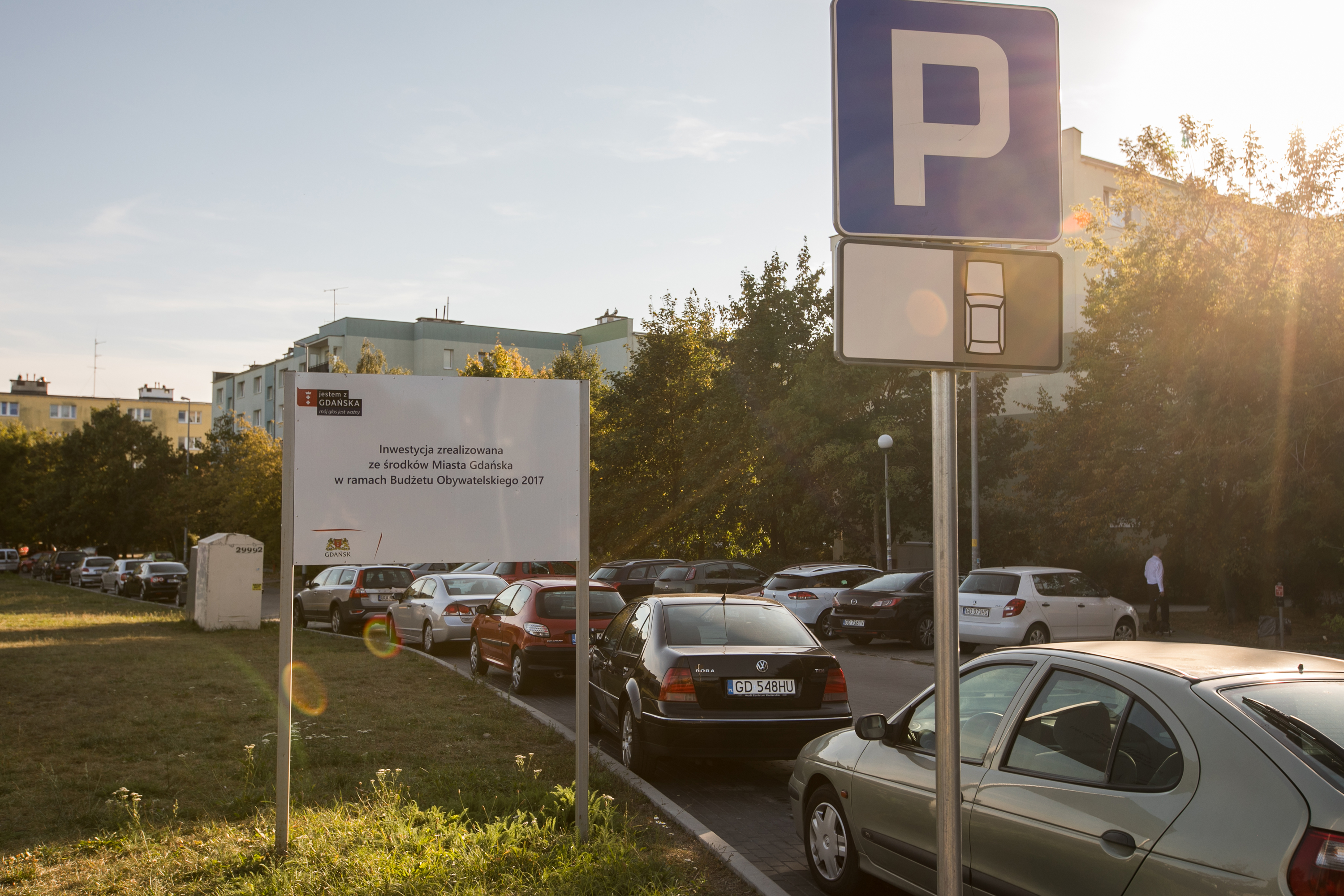 widok na zaparkowane samochody, znak drogowy informujący o miejscach parkingowych oraz tablica informująca o wykonanej inwestycji w ramch Budżetu Obywatelskiego 2017
