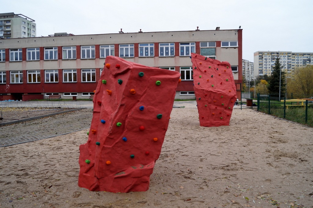 dwie czerwone ścianki wspinaczkowe na piasku, w tle budynek szkoły